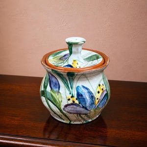 Può includere: Vaso in ceramica dipinto a mano con coperchio, con motivi floreali blu, verdi e gialli. Il vaso ha una forma arrotondata, un bordo marrone e un pomello bianco sul coperchio. Lo sfondo è rosa tenue.