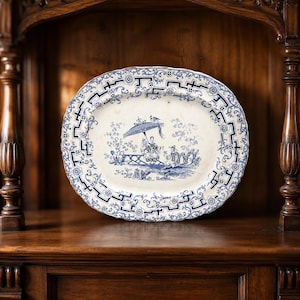 May include: An oval ceramic platter featuring a blue and white transferware pattern. The centre depicts a scene with figures under a parasol, surrounded by a decorative border of geometric lines and floral motifs on a cream-coloured background.