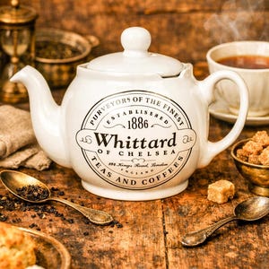 Op de afbeelding: Een witte keramische theepot met deksel, met de tekst "Whittard of Chelsea, Teas and Coffees, Established 1886". De afbeelding bevat een kopje thee, suikerklontjes en vintage lepels op een houten oppervlak.