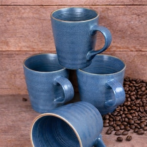 May include: Four blue ceramic coffee mugs with a textured finish and a light brown rim are stacked on a wooden surface. Coffee beans are scattered to the side. The mugs have curved handles.
