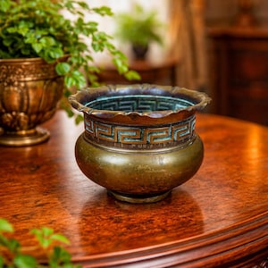 May include: An antique bronze planter with a fluted rim and a Greek key design. The planter sits on a dark wooden table. The planter's aged patina gives it a vintage aesthetic. A second bronze planter is visible in the background.