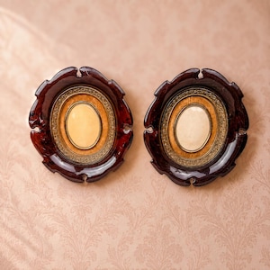 May include: Two matching oval picture frames featuring a dark brown, glossy, scalloped outer border. Each frame contains a central cream-colored oval insert surrounded by a decorative gold-toned metal trim with intricate embossed detailing.