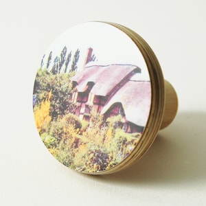 May include: A round wooden knob with a printed image of a thatched-roof cottage surrounded by greenery. The knob is a light brown color and has a smooth, polished finish.
