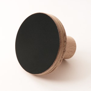 以下が含まれることがあります： 丸い黒い表面を持つ木製壁掛けフック。フックは自然な木製の仕上げの合板で作られています。