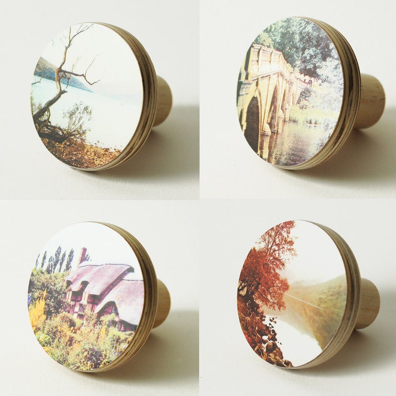 May include: Four wooden wall hooks with different scenic images. Each hook features a circular wooden base with a printed image of a landscape scene. The images include a lake with a tree, a bridge over a river, a cottage with a garden, and a foggy forest.