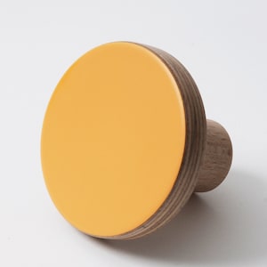 以下が含まれることがあります： 明るい黄色の前面を備えた丸い木製の壁掛けフック。フックは天然木目のエッジと、吊り下げ用の小さな円筒形の木製ペグを備えています。モダンなデザインです。