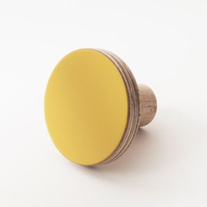 May include: A yellow wooden knob with a round, flat top. The knob is made of plywood and has a natural wood finish.