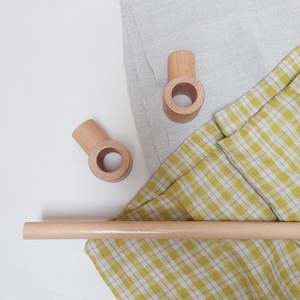 Peut inclure: Deux petits supports de tringle à rideaux en bois clair et une tringle en bois sont présentés sur un fond de tissu de lin gris et de tissu à carreaux jaune et blanc. Les supports ont une ouverture circulaire.