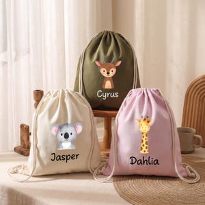 Könnte beinhalten: Drei Kordelzugbeutel in Beige, Olivgrün und Rosa. Jeder Beutel zeigt eine Aquarell-Tierillustration: einen Koala, ein Reh und eine Giraffe. Die Beutel sind mit den Namen personalisiert: Jasper, Cyrus und Dahlia.