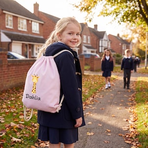 Könnte beinhalten: Ein hellrosa Kordelzugrucksack mit einem Giraffenmotiv und dem Namen "Dahlia" in schwarzer Schrift. Der Rucksack wird von einem jungen Mädchen in einem marineblauen Mantel und einer Schuluniform getragen. Andere Kinder gehen im Hintergrund auf einem Weg.