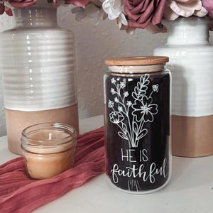 May include: A glass jar with a wooden lid and a black background. The jar has a white floral design and the text "He is faithful" written on it.