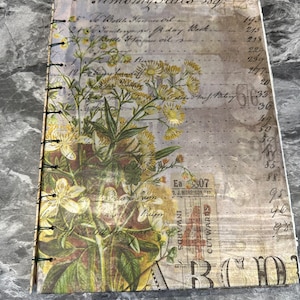 Op de afbeelding: Een vintage-stijl notitieboek met een bloemenontwerp met gele bloemen en groene bladeren. Het notitieboek heeft een bruine omslag met een witte achtergrond en een handgeschreven lijst met items en prijzen. Het notitieboek is gebonden met metalen ringen.