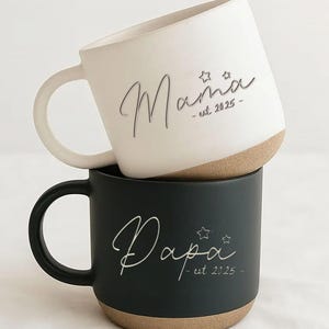 May include: Two ceramic mugs, one white and one black, stacked. The white mug has the word "Mama" in cursive with stars and "est. 2025". The black mug has "Papa" with stars and "est. 2025". Both mugs have a beige base.