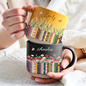 May include: Two ceramic mugs featuring personalised names and book-themed designs. The top yellow mug displays the name Emily with lily of the valley flowers and books. The bottom black mug shows the name Amelia with carnations and books.