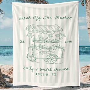 May include: Beach towel with a green and white striped background. The towel features a green illustration of a flower cart with the text "Fresh Off The Market" and "Emily's bridal shower Austin, TX".