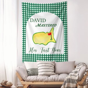 May include: A white tapestry with a green gingham border and the text "DAVID MASTERED His First Year." The design includes a yellow golf course map with a red flag. The tapestry is hanging above a wooden sofa with pillows.
