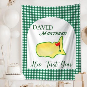 May include: A white banner with green gingham border and a central design. The design features a yellow golf course map with a red flag, and the text "DAVID MASTERED His First Year" in green script. The banner is likely for a golf-themed event.