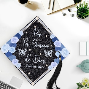 May include: A black graduation cap decorated with blue and white paper flowers, butterflies, and the text "Por la Gracia de Dios" and "Psalms 46:5". The cap has a black tassel and is trimmed with pearls.