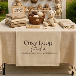 May include: A display of handmade knitwear and winter essentials. The table features a basket of yarn, crocheted teddy bears, folded blankets, and a sign that reads "Handmade with Love." The table runner displays the text "Cozy Loop Studio."