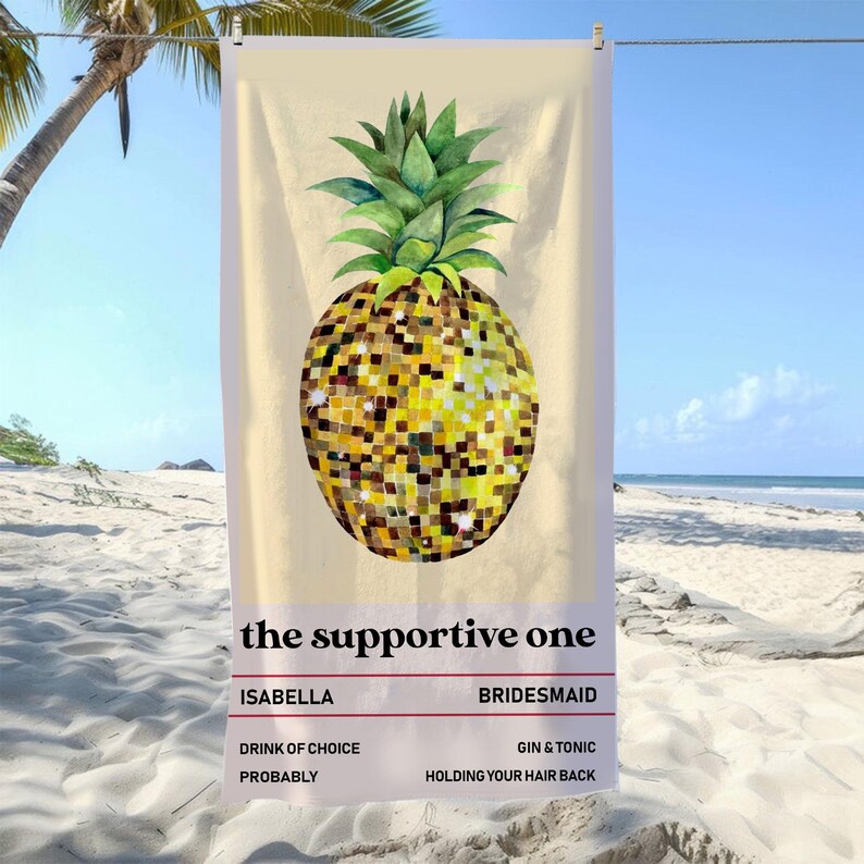 May include: Beach towel featuring a large pineapple design with a mosaic pattern in shades of yellow and gold. The pineapple has green leaves. Text at the bottom reads "the supportive one" with additional text below. The towel is hanging on a clothesline on a beach.