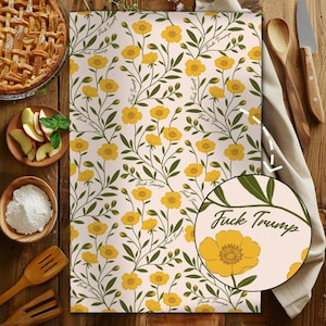 May include: A cream-coloured kitchen towel featuring a repeating floral pattern of yellow flowers and green vines. The words "Fuck Trump" are printed throughout the design. Cooking utensils and ingredients surround the towel.