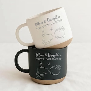 May include: Two ceramic mugs, one white and one black, stacked. Both mugs have the text "Mom & Daughter FOREVER LINKED TOGETHER" with floral and butterfly designs. The names "Olivia" and "Jennifer" are also included.
