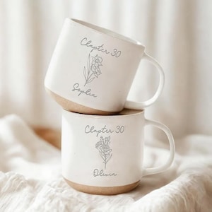 May include: Two white ceramic mugs stacked. Each mug has a light brown base and a curved handle. The text "Chapter 30" is above a floral illustration and a name on each mug. The mugs are on a white cloth.