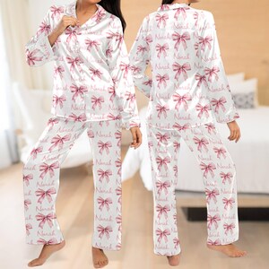 May include: White satin pyjamas with long sleeves and trousers. The pyjamas have a pink bow pattern with the name "Norah" repeated throughout.