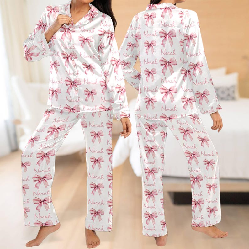 May include: White satin pyjamas with long sleeves and trousers. The pyjamas have a pink bow pattern with the name "Norah" repeated throughout.