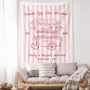 May include: A pink and white striped tapestry with a red illustration of a flower cart. The text "Fresh Off The Market" and "Emily's bridal shower AUSTIN, TX" are displayed. The tapestry is hanging above a wooden sofa with pillows.