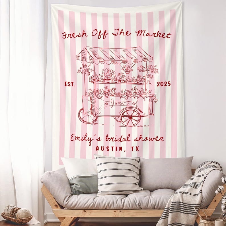 May include: A pink and white striped tapestry with a red illustration of a flower cart. The text "Fresh Off The Market" and "Emily's bridal shower AUSTIN, TX" are displayed. The tapestry is hanging above a wooden sofa with pillows.