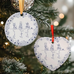 May include: White ceramic Christmas ornaments with blue floral borders. One is round, the other heart-shaped. Each features three cat illustrations with names "Max", "Lucky", and "Lila". Gold and red ribbons are attached for hanging.