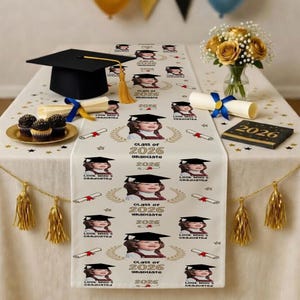 May include: A graduation-themed table setting with a black mortarboard, cupcakes, and a floral arrangement. A table runner features a repeating pattern of a child in a graduation cap, the text "Class of 2026 Graduate", and diploma illustrations.