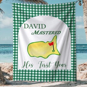 May include: A white beach towel with a green gingham border. The center features a golf course design with the text "DAVID MASTERED His First Year". The golf course is yellow with a red flag.