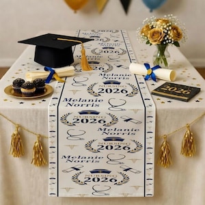 May include: A graduation table setting with a black mortarboard, cupcakes, and a floral arrangement. A white table runner features "Nursing 2026" and the name "Melanie Norris" in navy and gold. Decorative tassels and a small book complete the scene.