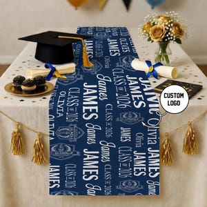 May include: A navy blue graduation table runner with white text that reads "James", "Olivia", and "Class of 2026". The runner is decorated with graduation caps, diplomas, and a bouquet of yellow roses.