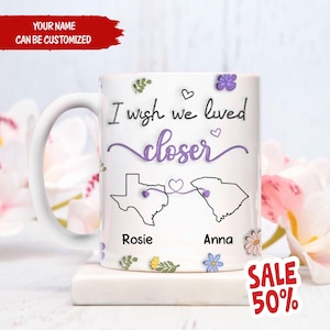May include: White ceramic mug with a floral design and the text "I wish we lived closer" with outlines of Texas and South Carolina connected by a heart string. The names "Rosie" and "Anna" are written below the states.