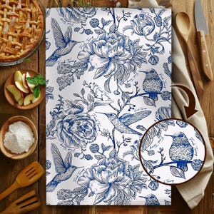 Peut inclure: Torchon de cuisine blanc avec un motif floral et colibri bleu. Le torchon présente une illustration détaillée de fleurs et de colibris, avec le texte "Fuck Trump" subtilement intégré dans le motif. Des articles de cuisine entourent le torchon.