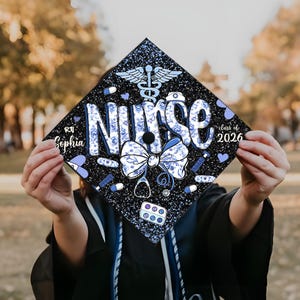 May include: A black graduation cap decorated with blue and white floral designs, medical symbols, and the word "Nurse." The cap also includes the name "Sophia" and the year "2026."
