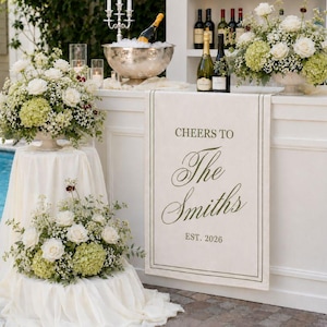 May include: A cream-coloured fabric banner featuring the dark green text "CHEERS TO The Smiths EST. 2026" framed by a thin border. The banner hangs on a white bar counter decorated with floral arrangements, a silver ice bucket, and wine bottles.