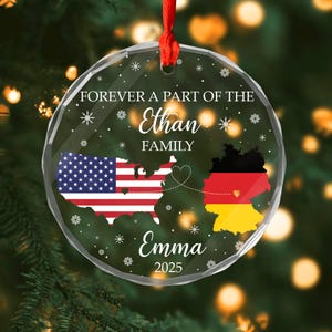 May include: Clear, round Christmas ornament with a red ribbon. The ornament features the text "Forever a part of the Ethan Family" and "Emma 2025". It includes the US and German flags connected by a heart.