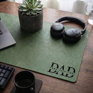 May include: A green desk mat with the word "DAD" and the names "EMMA * JACK * TOM" printed in black. A succulent in a gray pot, black headphones, a laptop, a keyboard, a black mug, and a coaster are on the wooden desk.