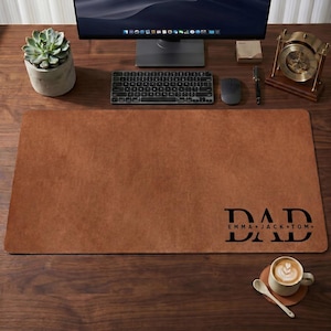 May include: A brown leather desk mat with the word "DAD" and the names "EMMA JACK TOM" printed in black. The mat is rectangular and sits on a wooden desk, with a keyboard, mouse, and monitor visible.