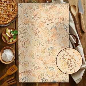 May include: A beige kitchen towel with a repeating pattern of outlined maple leaves in shades of brown, orange, and green. The towel is displayed on a wooden surface, alongside baking ingredients and utensils, suggesting a fall or autumn theme.