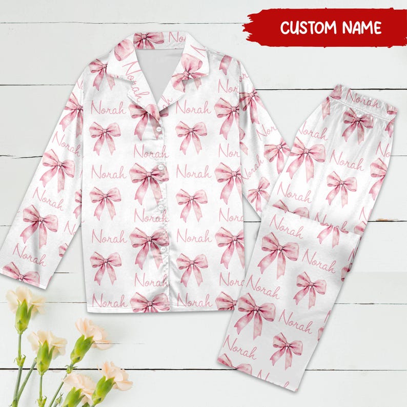May include: White pyjamas with long sleeves and trousers. The pyjamas have a pink bow pattern and the name "Norah" repeated throughout the fabric.