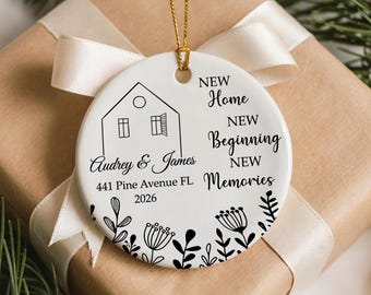 May include: A white ceramic ornament with a gold string for hanging. The ornament features a house illustration, the names "Audrey & James", address, and the year 2026. Text includes "New Home, New Beginning, New Memories". Black floral design at the bottom.