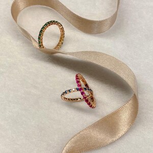 May include: Four gold rings with colorful gemstones. The rings are on a beige ribbon. The text "Argolla sin fin oro rosa" is at the bottom of the image.