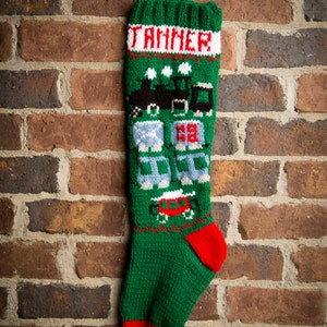 May include: Green knitted Christmas stocking with the name "TANNER" in white letters. The stocking features a train and house design in black, blue, red, and white. The toe is red, and the top cuff is green.