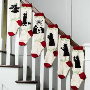 May include: A set of six Christmas stockings with a cream-colored base, red cuffs and heels, and green zigzag patterns. Each stocking features a black silhouette of a nativity scene or angel. The stockings are displayed on a staircase.