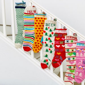 Könnte beinhalten: Sieben bunte, gestrickte Weihnachtsstrümpfe mit aufgestickten Namen, darunter "Isla", "Beck", "Emma", "Mama" und "Prisley". Die Strümpfe hängen an einem weißen Geländer.